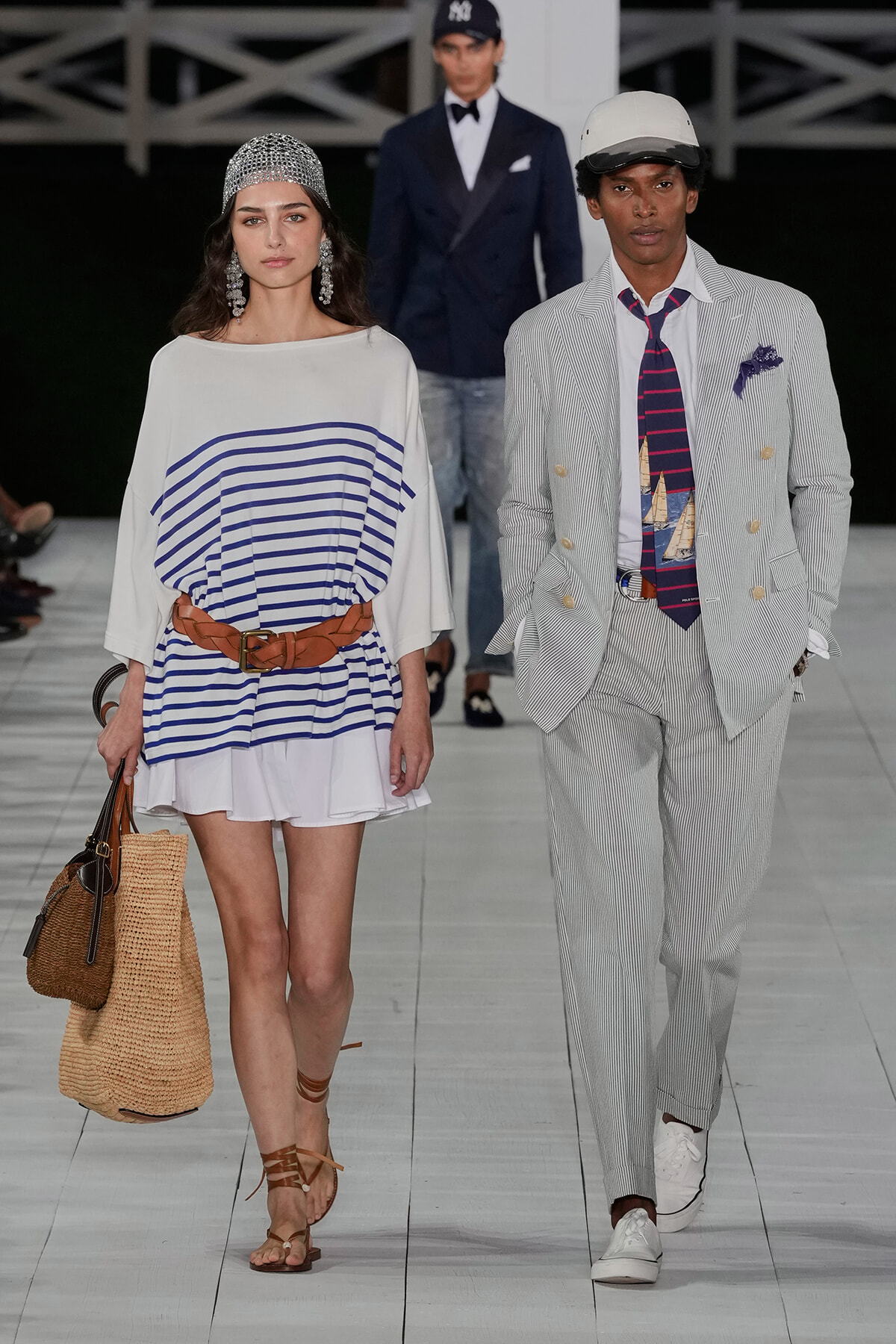 Two models walk a fashion runway: woman in a white and blue striped tunic with a wide brown belt, straw tote, and brown sandals; man in a light pinstripe suit with nautical tie and cap.