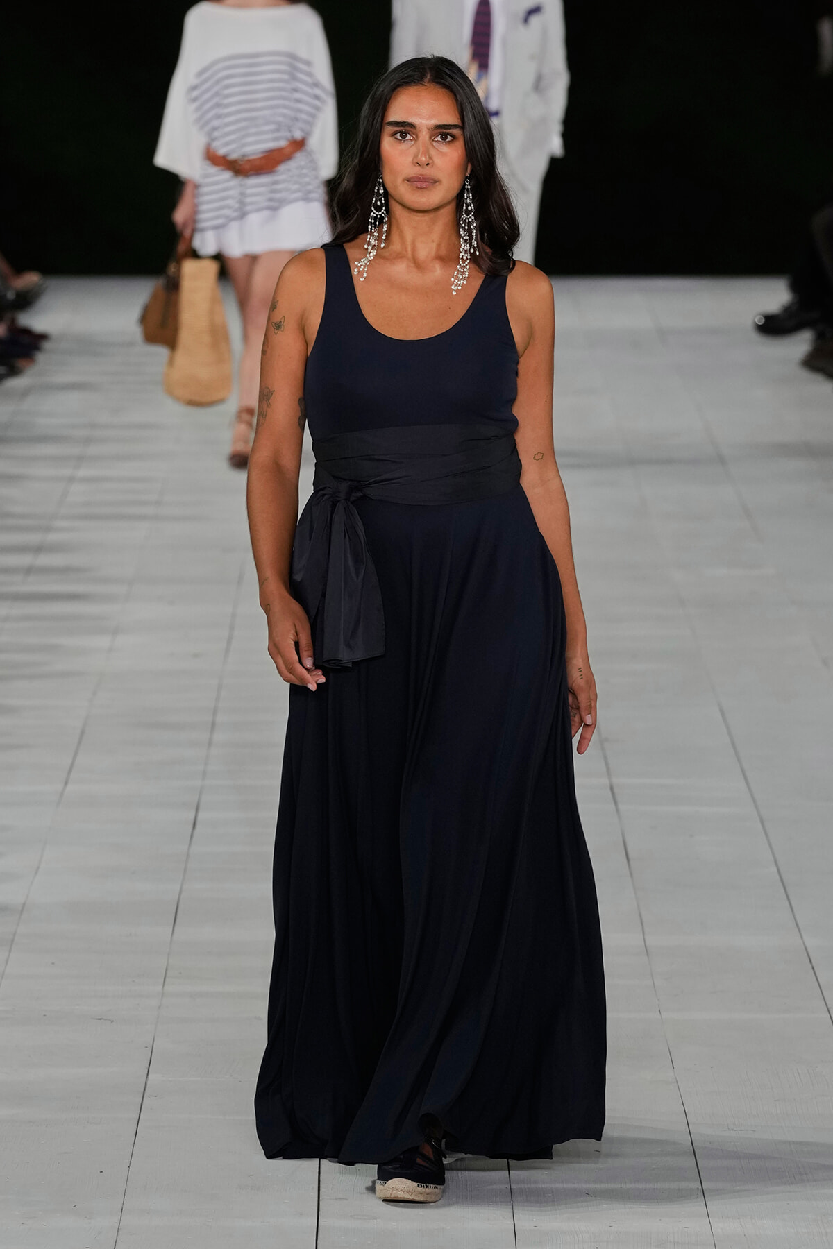 Model walks a navy sleeveless maxi dress with a wrapped waist sash and long chandelier earrings on a runway.