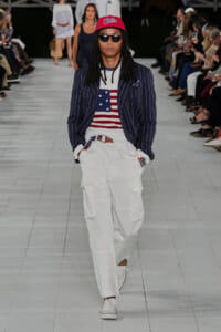 Male model walking a runway in a navy pinstripe blazer, American flag sweater, white cargo pants, red cap, and sunglasses.