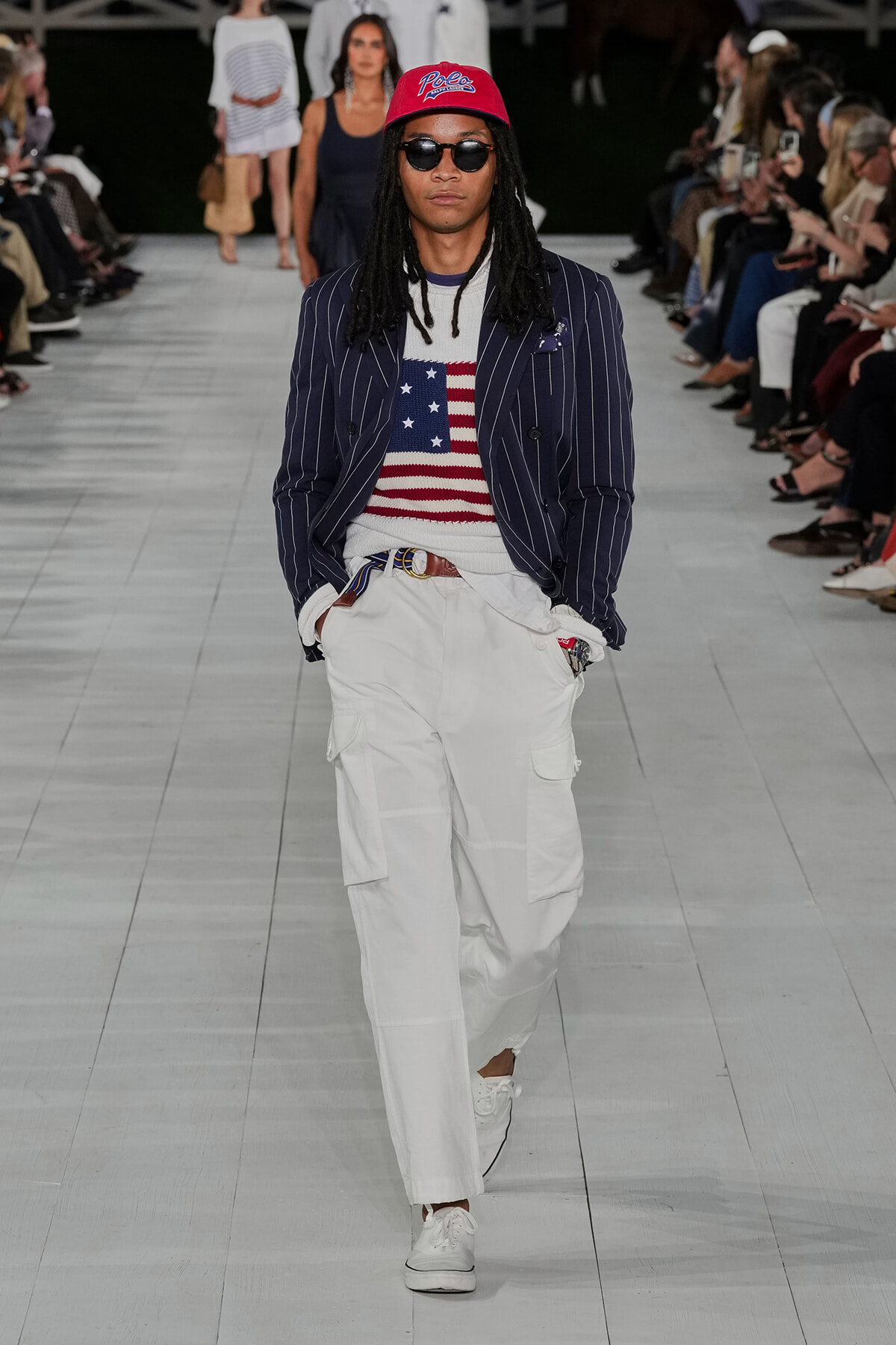 Male model walking a runway in a navy pinstripe blazer, American flag sweater, white cargo pants, red cap, and sunglasses.