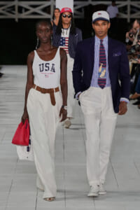 Two models walk a fashion runway in patriotic outfits: a woman in a white USA tank, wide white pants, brown belt, and red bag; a man in a navy blazer, striped shirt, nautical tie, white pants, and a cap.