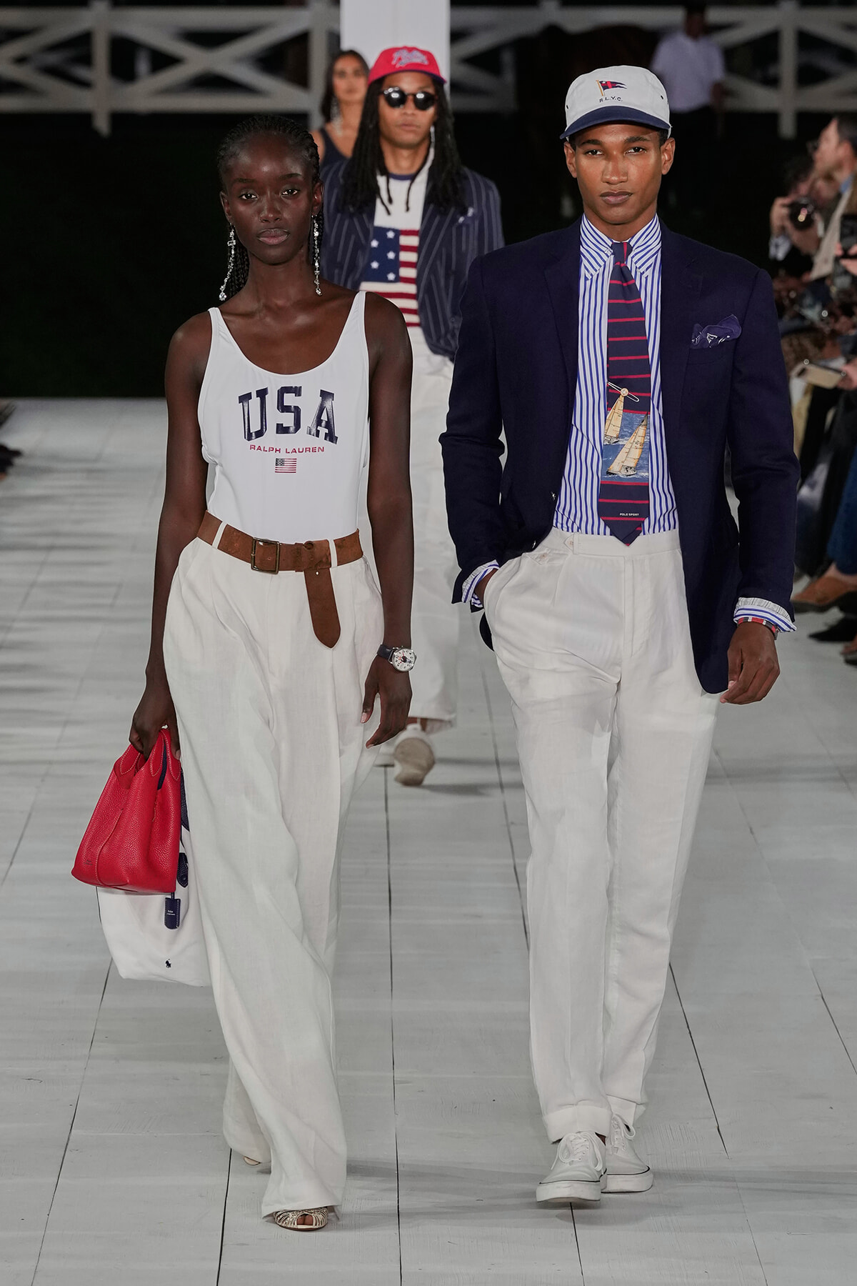 Two models walk a fashion runway in patriotic outfits: a woman in a white USA tank, wide white pants, brown belt, and red bag; a man in a navy blazer, striped shirt, nautical tie, white pants, and a cap.