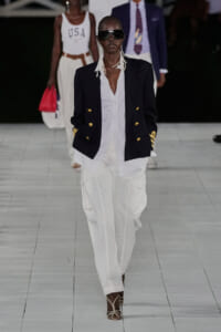 Model on a runway wearing a navy double-breasted blazer with gold buttons over a white shirt and white cargo pants, with oversized sunglasses.