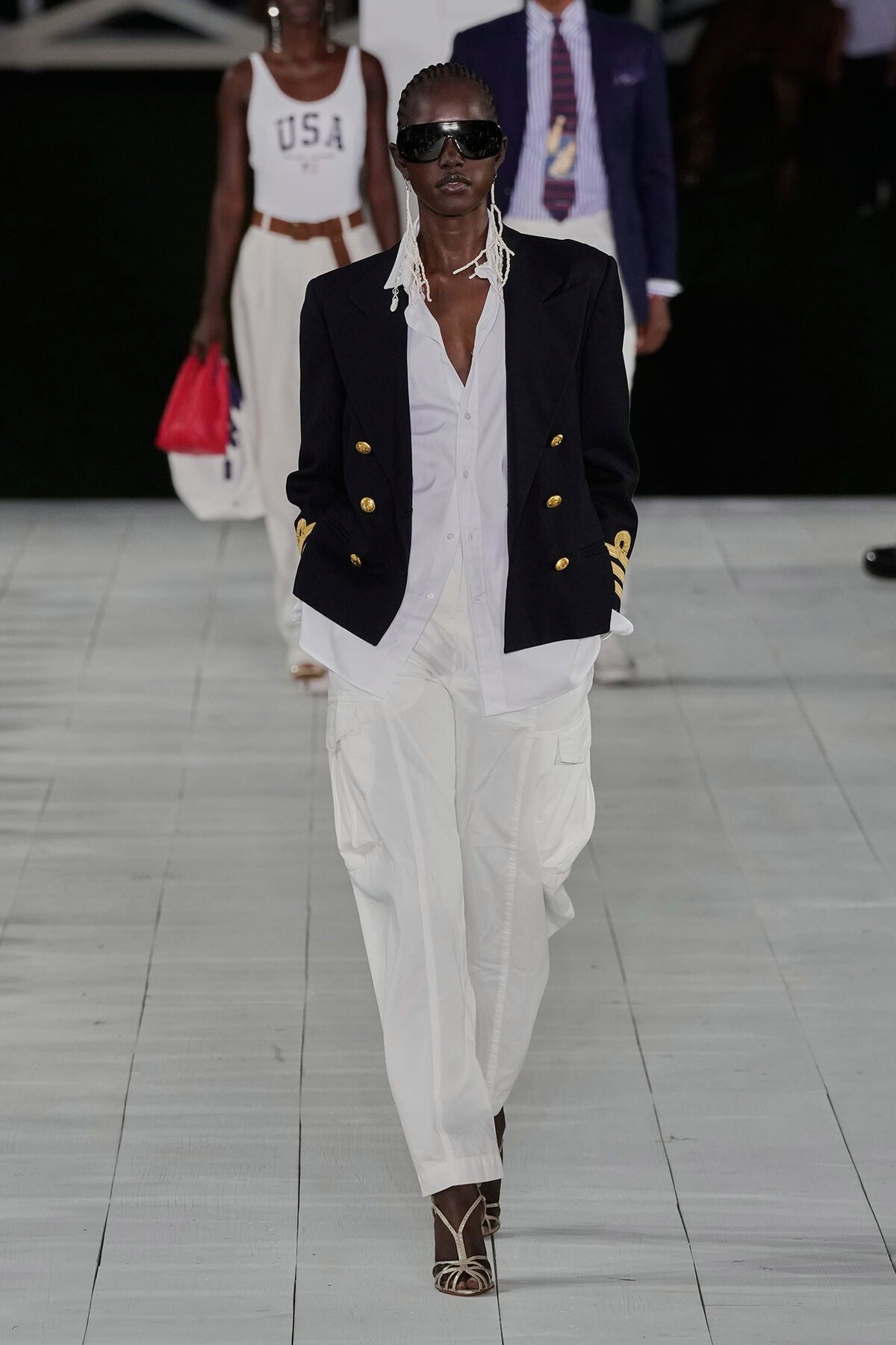 Model on a runway wearing a navy double-breasted blazer with gold buttons over a white shirt and white cargo pants, with oversized sunglasses.