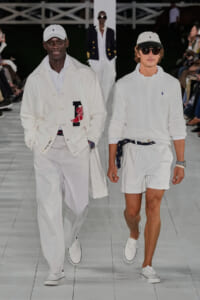 Two male models walk a fashion runway in all-white outfits: one in a white cap, long cardigan, and pants; the other in a white cable-knit sweater and shorts with a navy belt, sunglasses, and a cap.