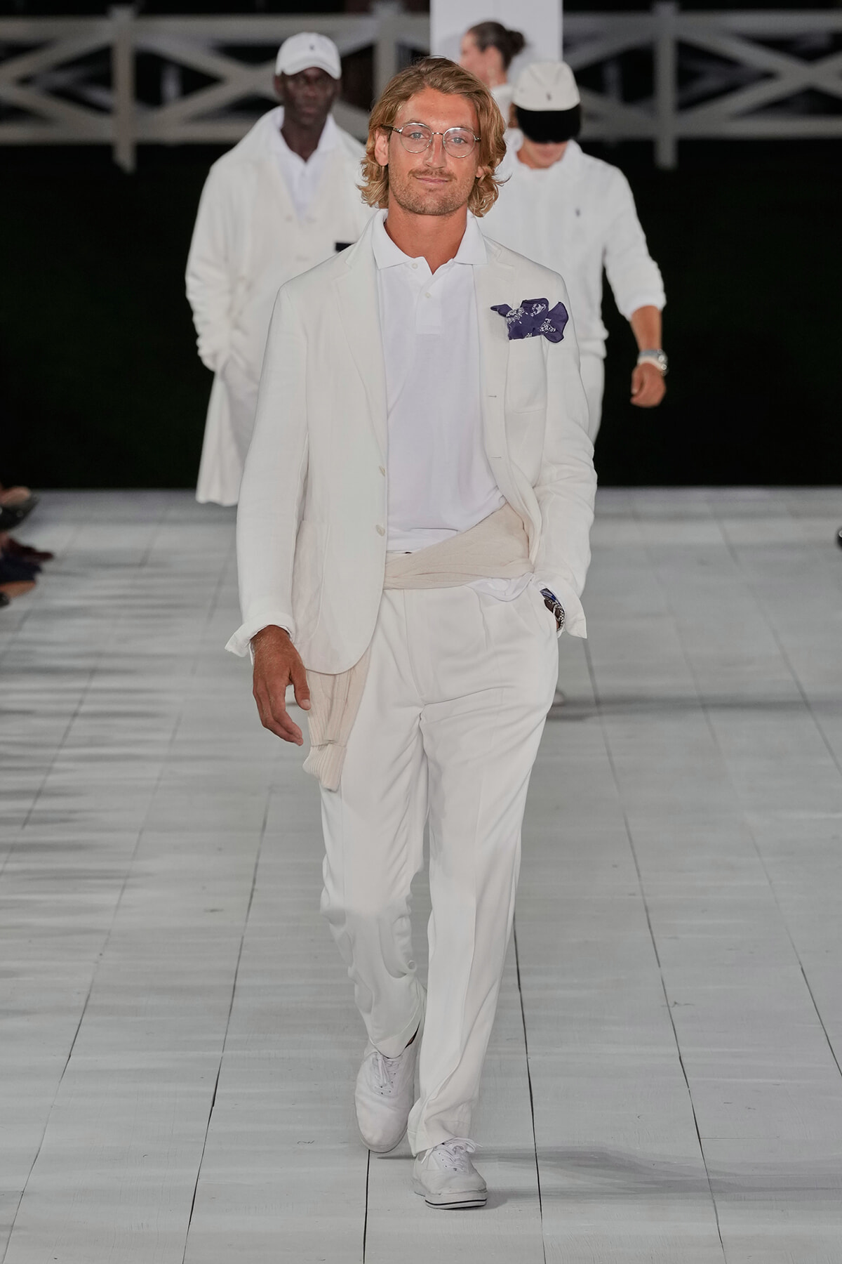 Male model walks the runway in an all-white outfit: blazer, polo, and trousers, with a beige sweater tied at the waist and a blue pocket square; white sneakers and background models in white.