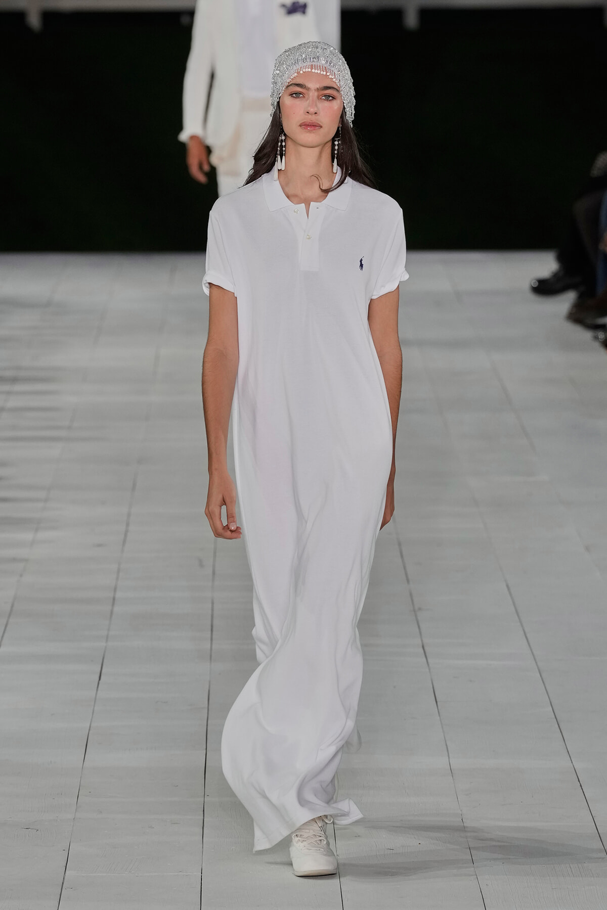 Model walking the runway in a long white polo-length dress with a small dark logo on the chest, paired with a sparkling beaded headpiece and dangling earrings.