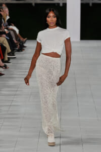 Model walking a fashion runway in a white knotted crop top and a long beaded white skirt, off-white wedges visible, audience along the sides.