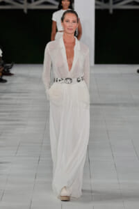 Model walking a runway in a sheer white blazer with deep V, cinched by a silver belt over a long flowing white skirt, wearing dangling earrings.