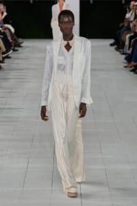 Model wearing an all-white textured suit with a long blazer, white vest, and wide-leg satin pants on a fashion runway, accessorized with a pearl collar necklace.