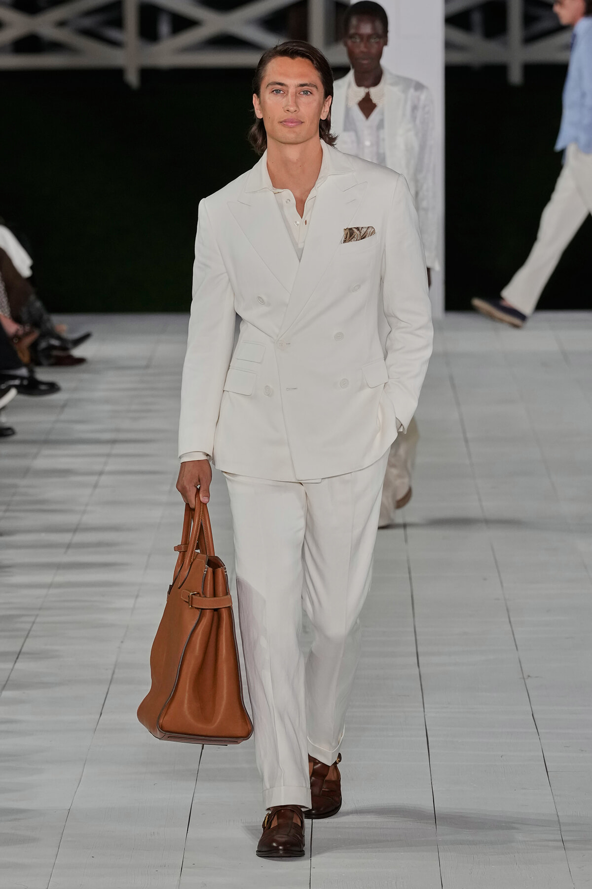 Male model wearing a white double-breasted suit with a pocket square, walking on a runway while carrying a brown leather tote bag.
