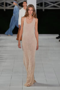 Runway model walking in a sheer nude-beige mesh full-length dress with a scoop neckline on a white runway.