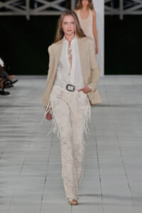 Fashion model on a runway wearing a beige blazer, white shirt, and cream embroidered fringe pants with a belt.