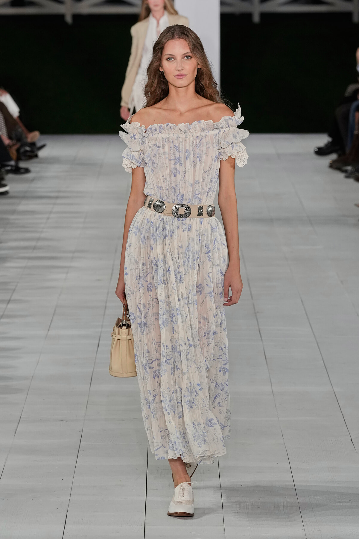 Model walking a runway in an off-shoulder, cream and pale blue floral maxi dress with ruffled neckline and lace sleeves, cinched with a decorative belt, carrying a beige handbag.
