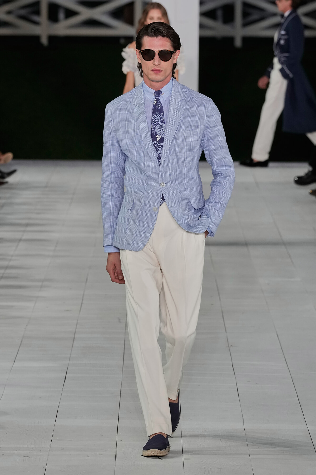 Male model in a light blue, checkered blazer with a blue patterned tie and white trousers on a fashion runway, wearing dark sunglasses.