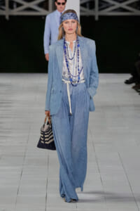 Model on a runway wearing a light denim blazer and wide-leg jeans with a striped shirt, blue beaded necklaces, and a woven tote bag.
