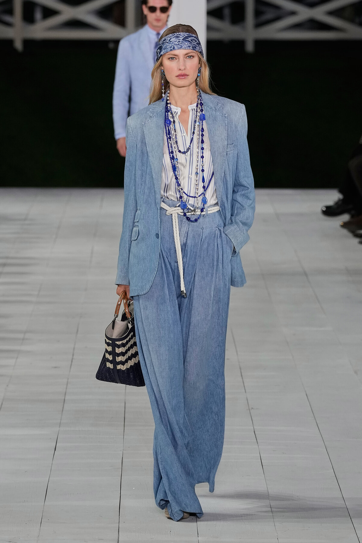 Model on a runway wearing a light denim blazer and wide-leg jeans with a striped shirt, blue beaded necklaces, and a woven tote bag.