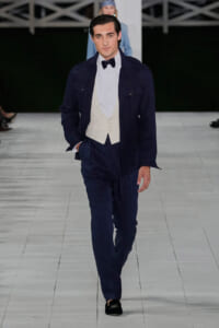Male model walks the runway in a navy suit with a cream waistcoat and bow tie, hands in pockets.