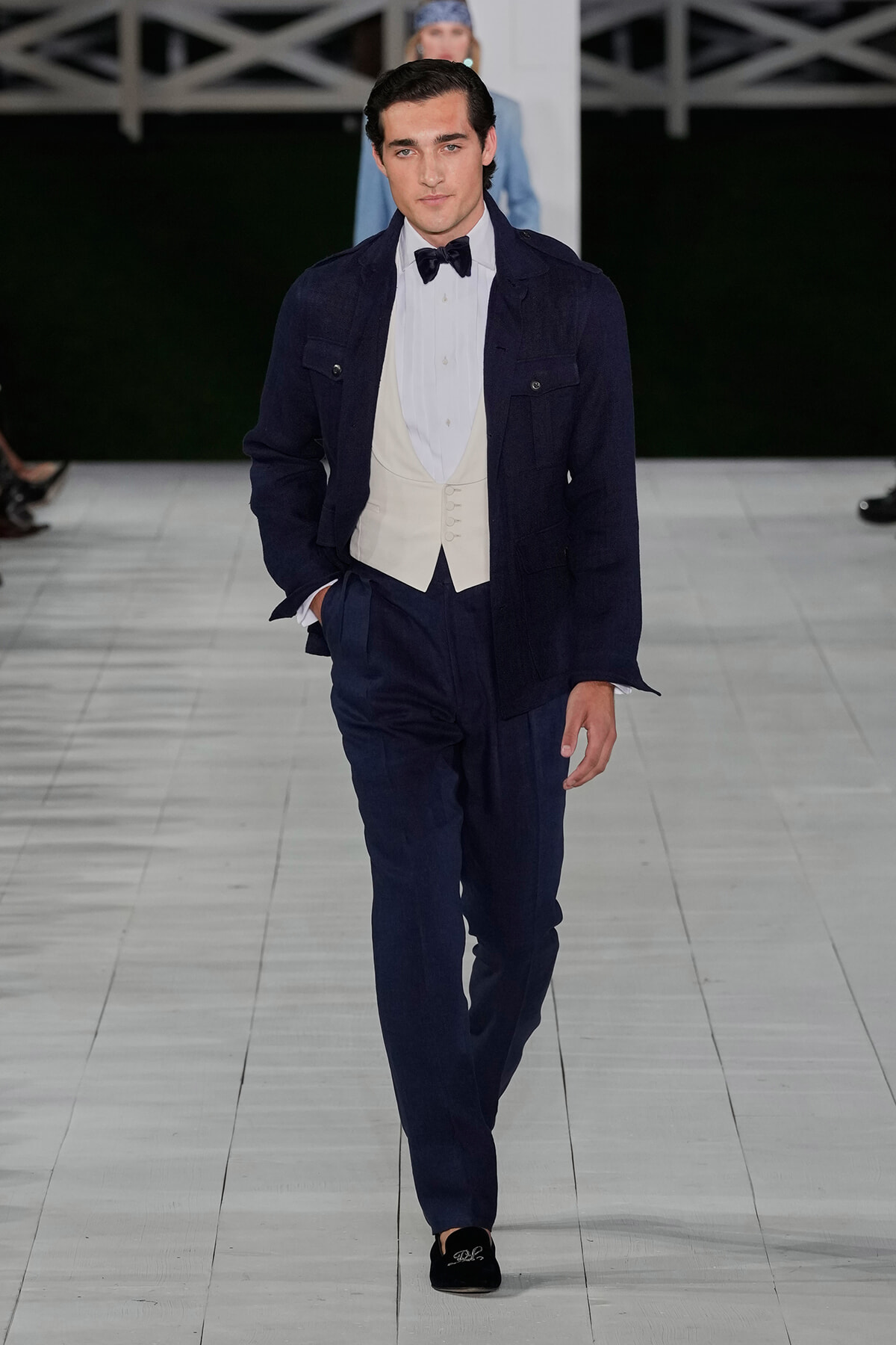 Male model walks the runway in a navy suit with a cream waistcoat and bow tie, hands in pockets.