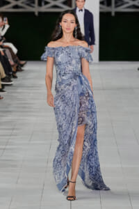 Model walks the runway in a blue off-shoulder lace gown with a side wrap and high slit, black strappy heels.
