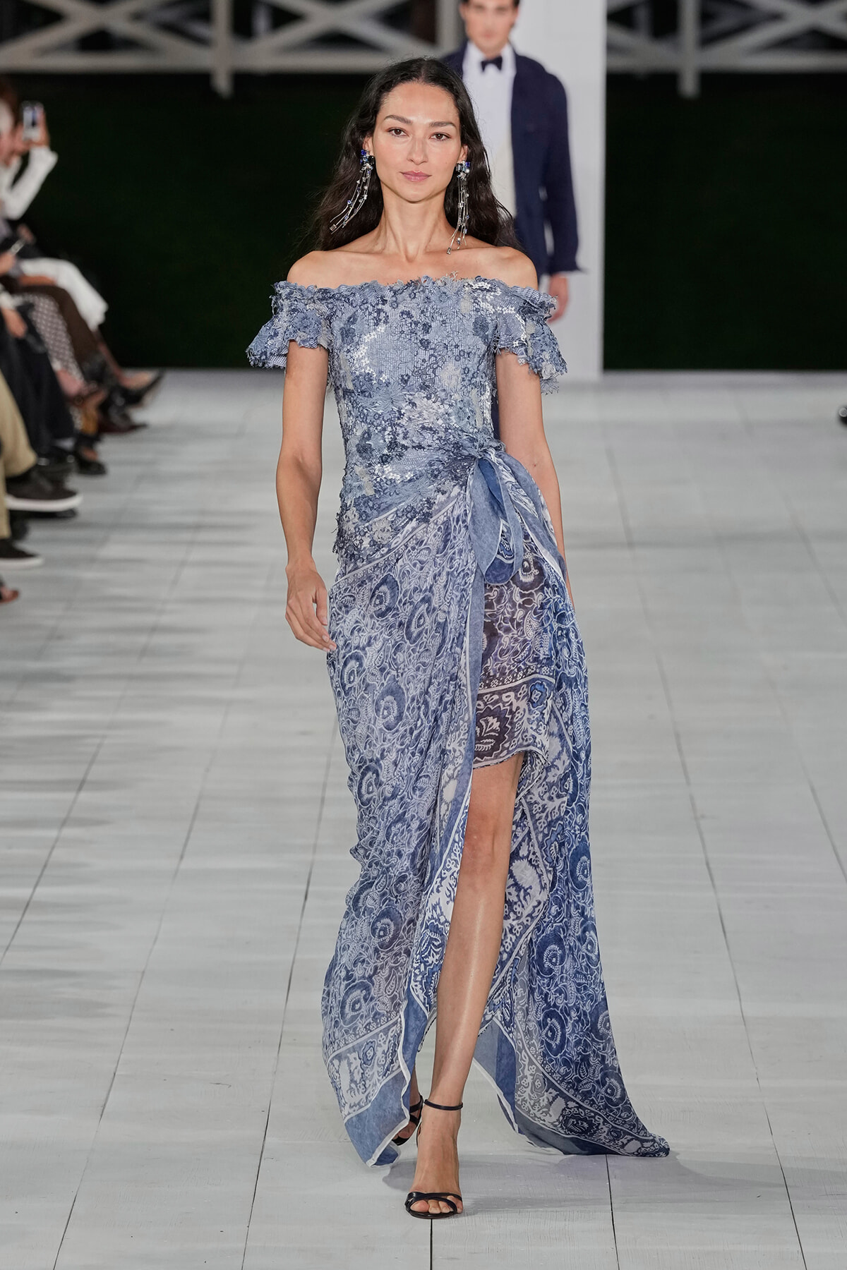 Model walks the runway in a blue off-shoulder lace gown with a side wrap and high slit, black strappy heels.