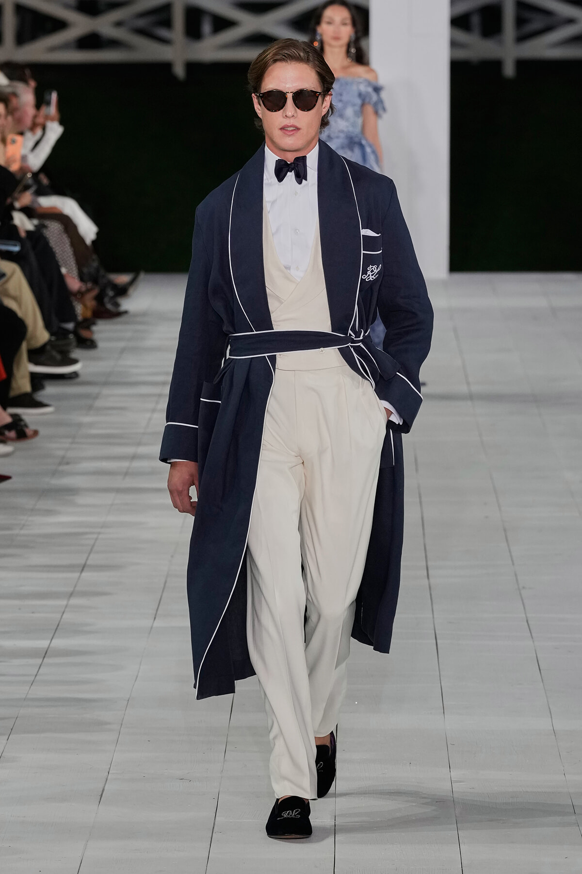 Male model on a runway wearing a navy robe with white piping over a cream suit, bow tie, and sunglasses.