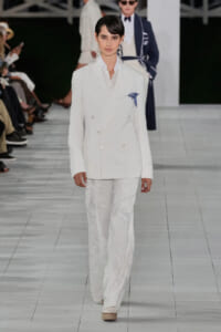 Model on a runway wearing a white double-breasted suit with embroidered detailing and a blue pocket square
