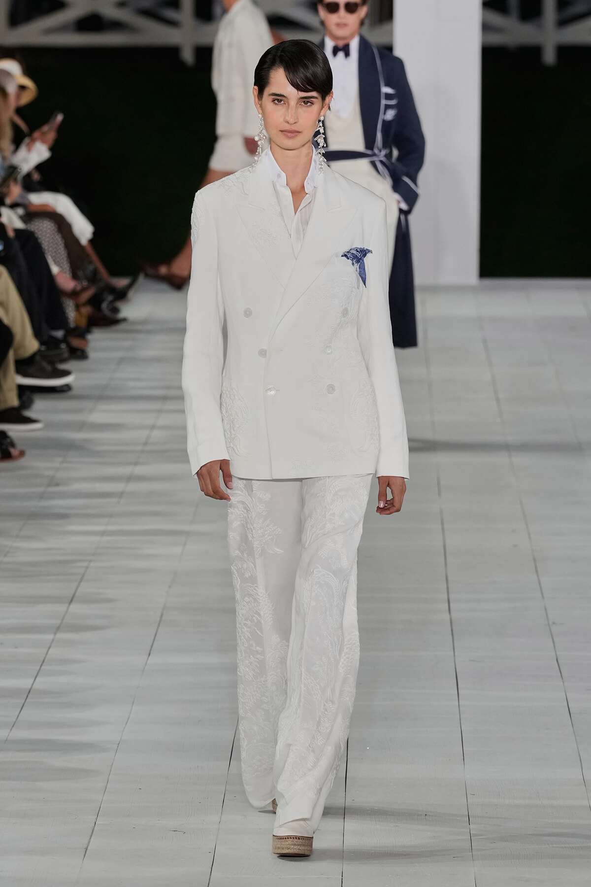 Model on a runway wearing a white double-breasted suit with embroidered detailing and a blue pocket square