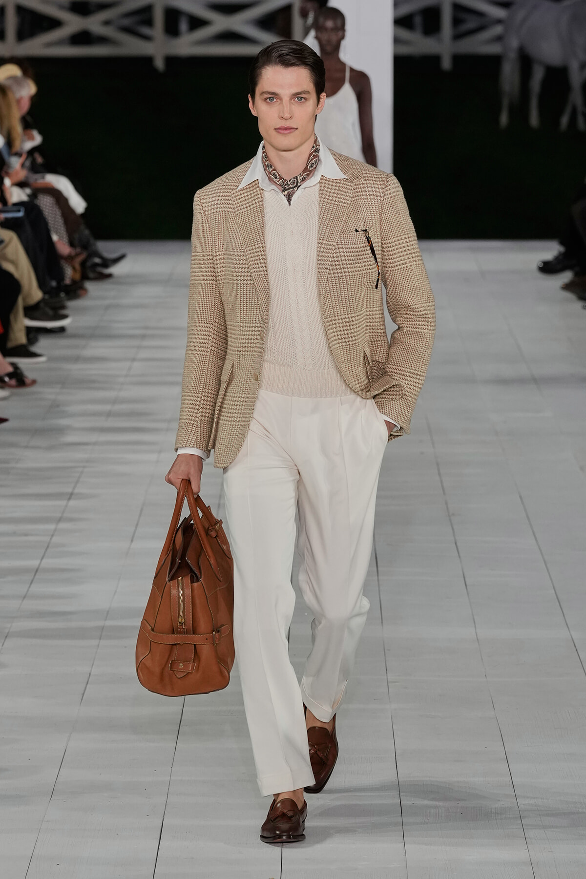 Male model on a runway wearing a tan plaid blazer, cream knit sweater, white trousers, brown loafers, and carrying a brown leather duffel bag.