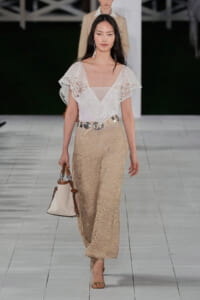 Model walks a runway in a white lace V-neck blouse and beige crocheted wide-leg pants with a light belt and taupe handbag.
