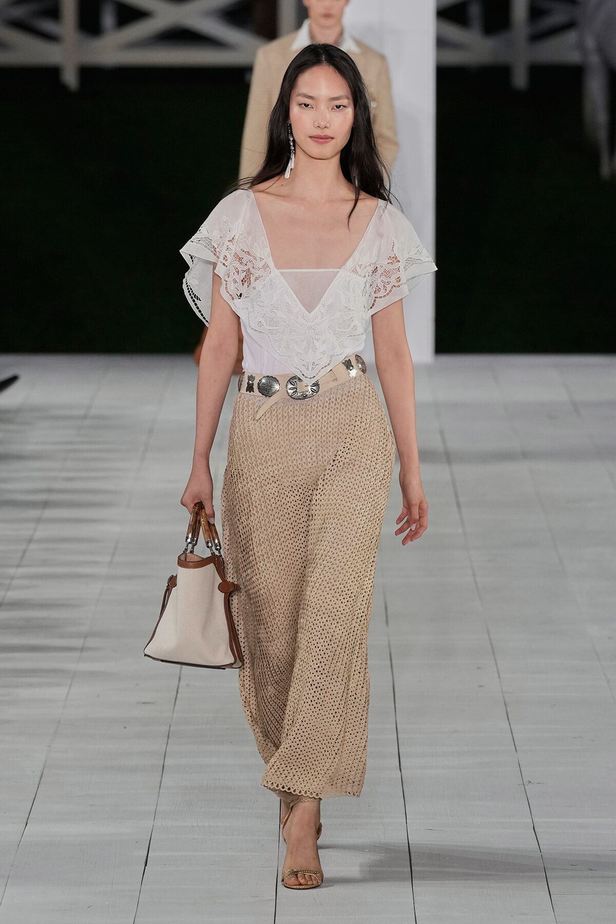 Model walks a runway in a white lace V-neck blouse and beige crocheted wide-leg pants with a light belt and taupe handbag.