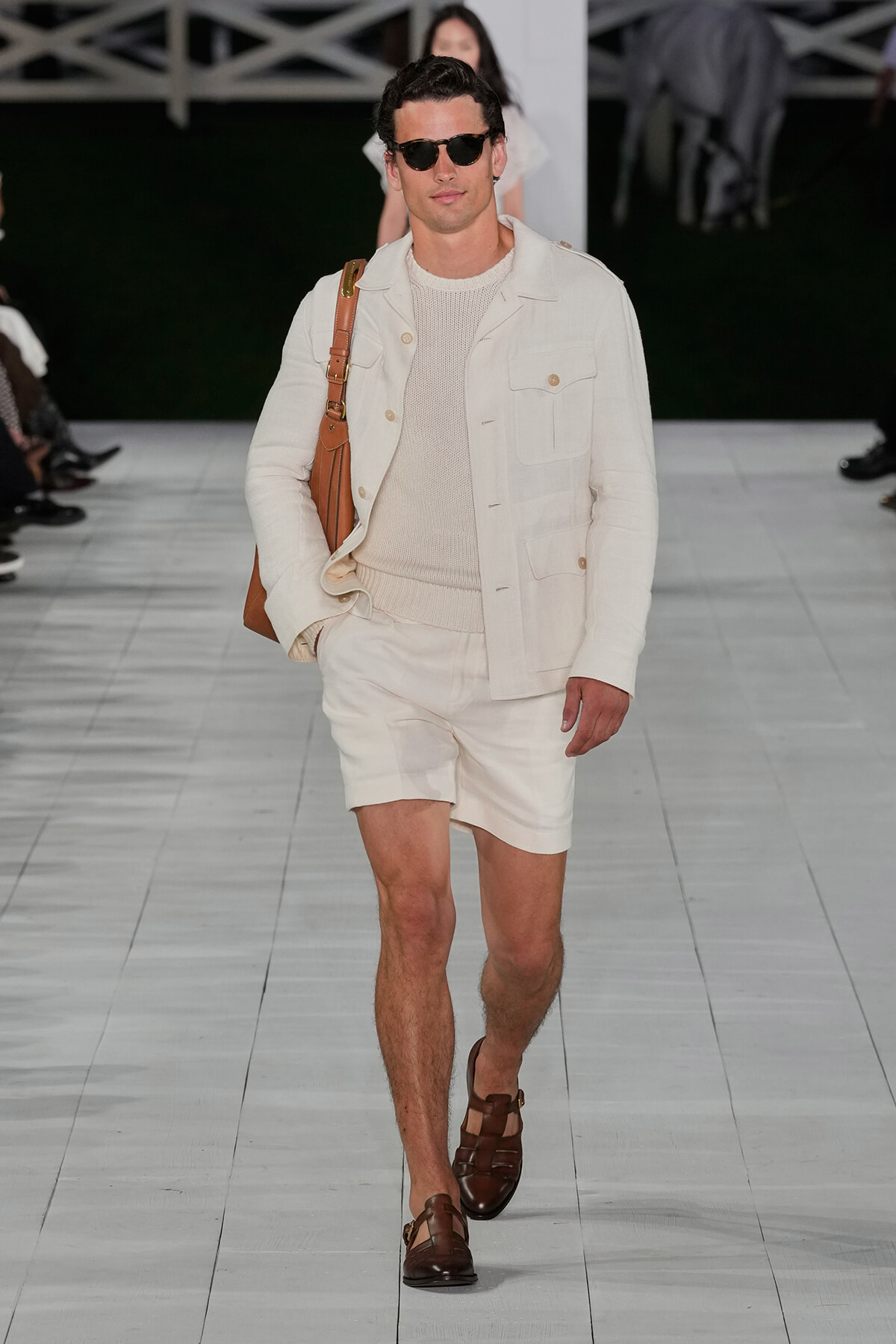 Male model on a fashion runway wearing an all‑cream outfit: knit sweater, utility jacket, matching shorts, brown sandals, dark sunglasses, and a brown shoulder bag.