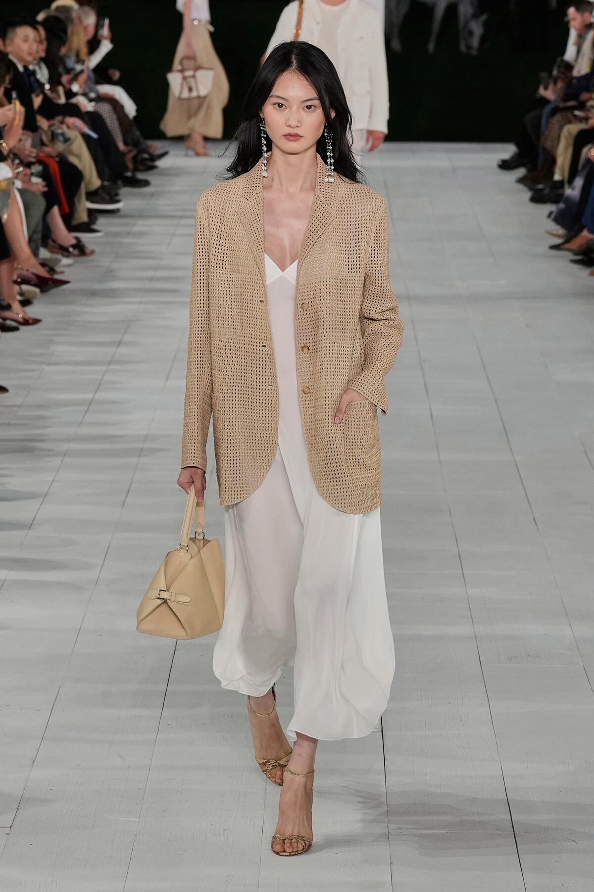 Model walking runway in a beige knit blazer over a white dress, carrying a beige handbag and wearing dangling earrings.
