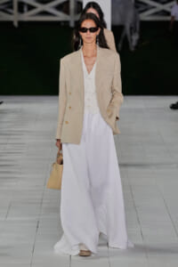 Model walks the runway in a beige oversized blazer over a white waistcoat and flowing white wide-leg trousers, with dark sunglasses, long dangling earrings, and a pale beige handbag.