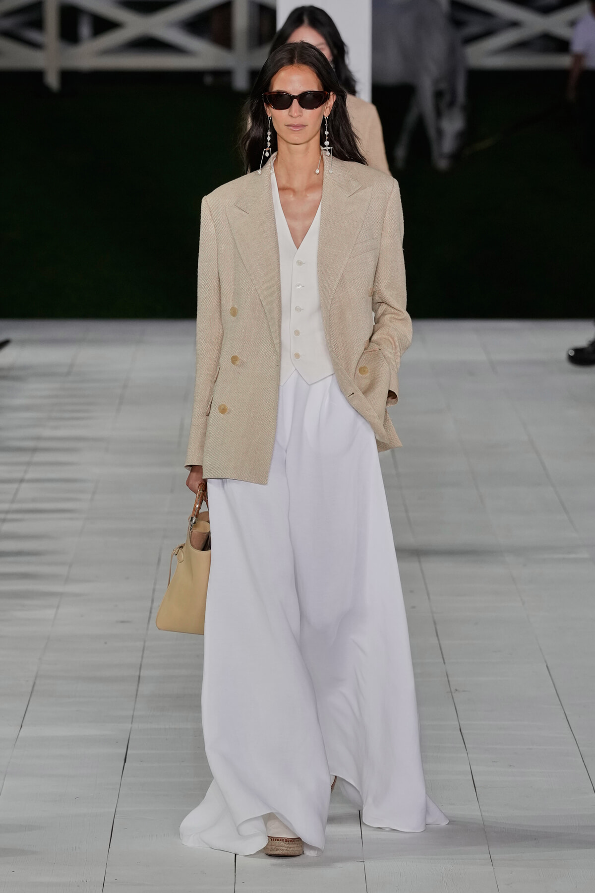 Model walks the runway in a beige oversized blazer over a white waistcoat and flowing white wide-leg trousers, with dark sunglasses, long dangling earrings, and a pale beige handbag.