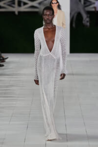 Model on a fashion runway wearing a long white crochet gown with a deep V-neck and long sleeves, accessorized with dangling earrings.