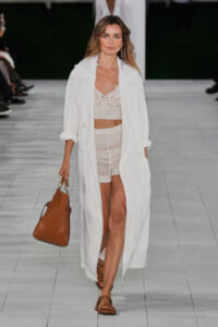 Model walking a fashion show in a long white coat worn over a white lace bralette and crochet shorts, carrying a brown handbag.