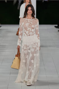 Model on a runway wearing a full-length white floral lace gown with sheer mesh, carrying a tan straw tote.