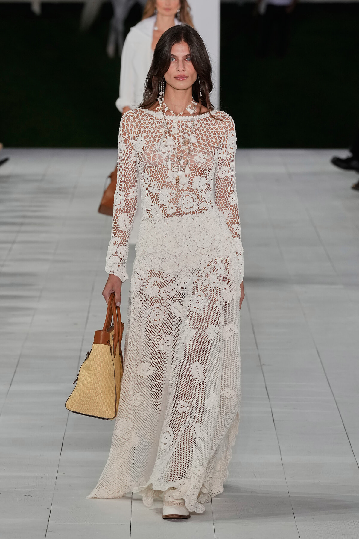 Model on a runway wearing a full-length white floral lace gown with sheer mesh, carrying a tan straw tote.