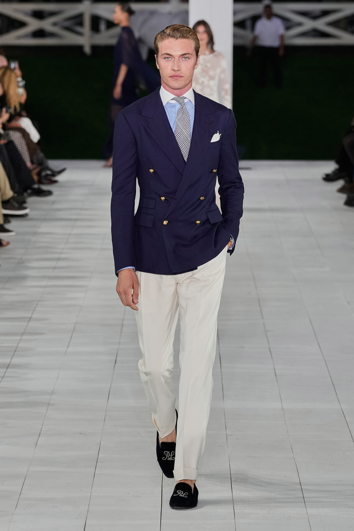 Male model walking a fashion runway in a navy double-breasted blazer, light blue shirt, patterned tie, white trousers, and black embroidered loafers.