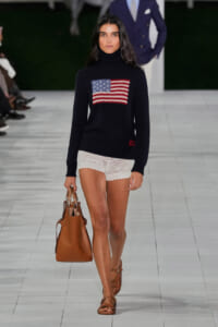 Model walking a runway in a black turtleneck sweater with an American flag patch, white crochet shorts, and brown handbag.