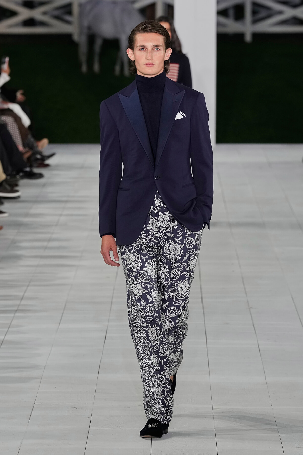 Male model walking a runway in a dark navy blazer, black turtleneck, white pocket square, and navy-and-white paisley print trousers.