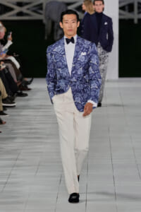 Male model walking a runway in a blue paisley blazer, white shirt, black bow tie, and white trousers.
