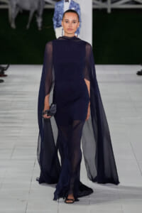 Model in a navy sheer gown with a flowing cape, walking the runway holding a small black clutch.