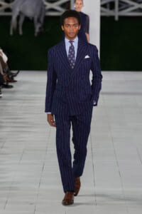 Male model walking a runway in a navy pinstripe double-breasted suit, white shirt, patterned tie, brown oxford shoes, and pocket square