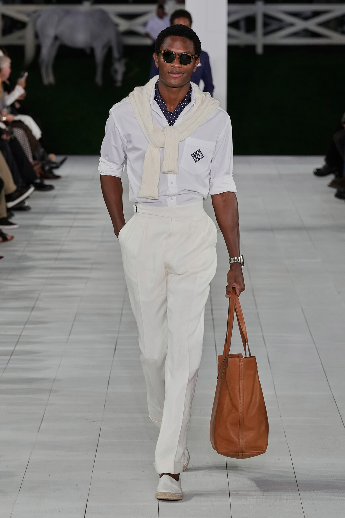 Male model walking runway in all-white outfit: white shirt with logo, cream sweater draped over shoulders, white trousers, and a large tan tote bag.