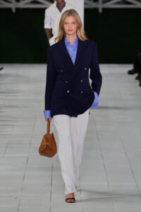 Model in a navy double-breasted blazer over a light blue shirt, white trousers, black sandals, carrying a brown handbag.