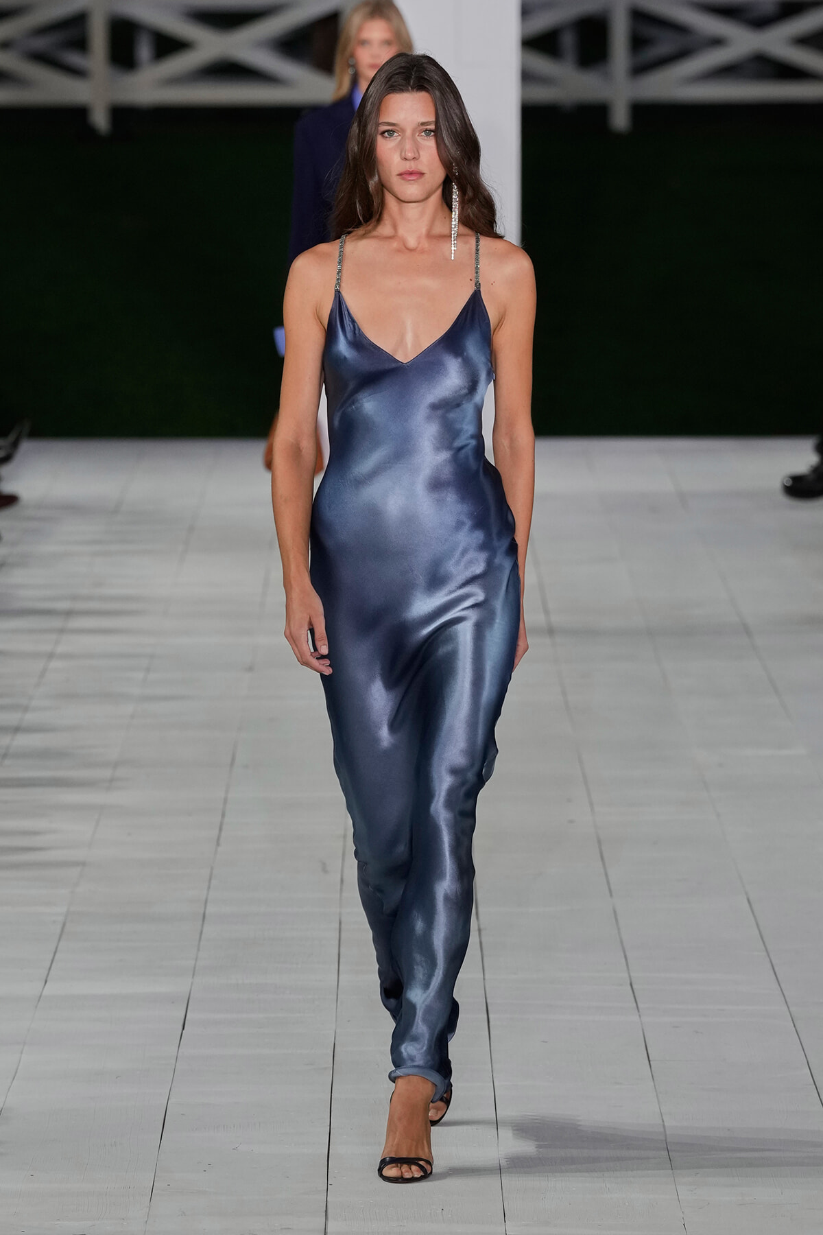 Model walking a runway in a satin navy slip dress with thin straps and a V-neck, wearing a long dangling earring and black heeled sandals