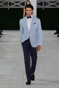 Male model on a runway wearing a light blue blazer, white shirt, navy trousers, black bow tie, pocket square, and loafers.