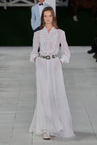 Model on a runway wearing a sheer white maxi dress with lace bib, belted at the waist with a large silver belt, hands in pockets.